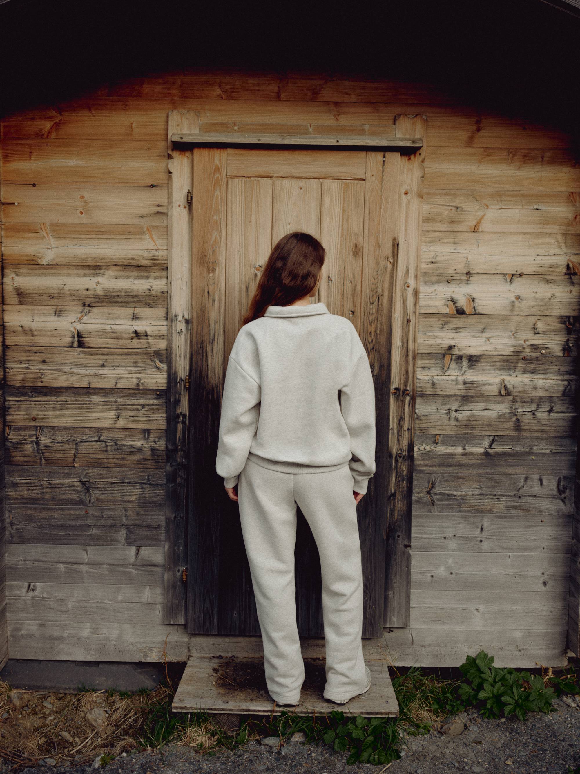 Woman wearing a grey melange CASPARA CS rugby shirt facing a rustic wooden cabin door.